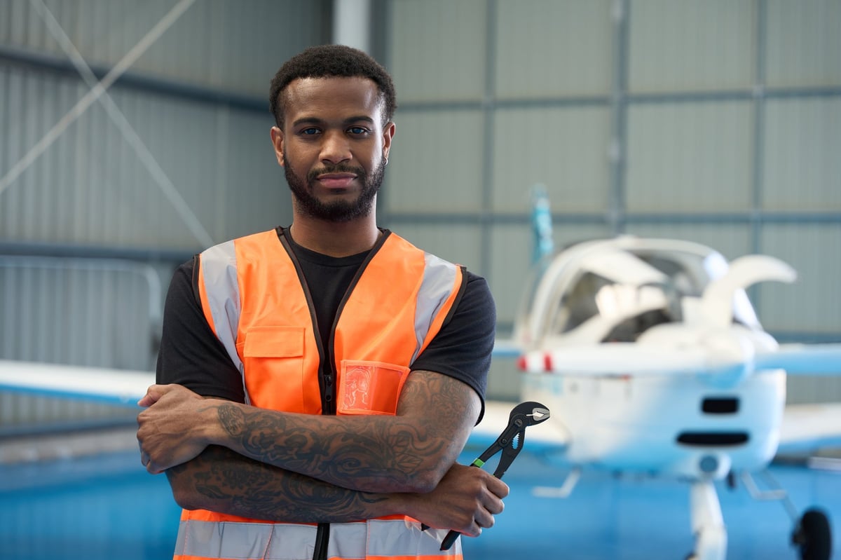 Confident aircraft mechanic holding pliers while posing in hangar with small passenger airplane in background