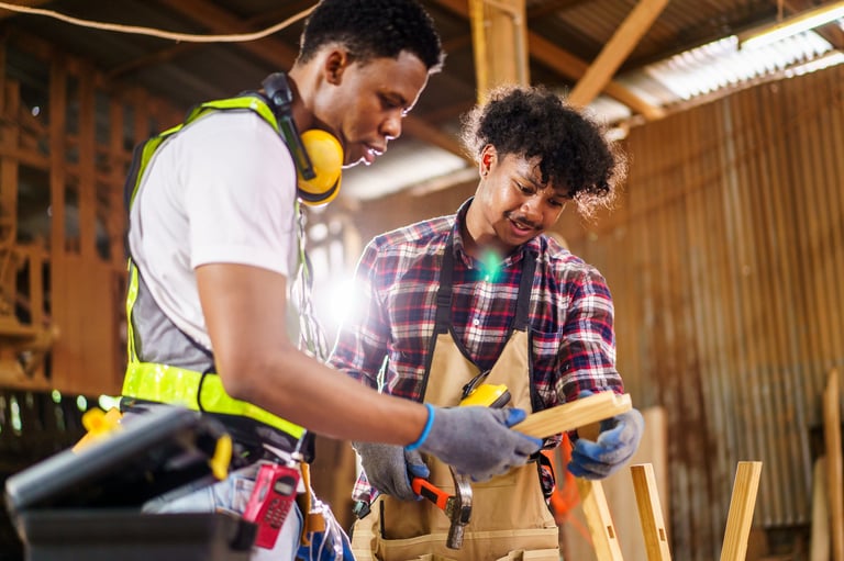 Two African carpenters collaborate seriously in a woodworking shop, discussing design plans for their current project. Scene highlights teamwork, craftsmanship, and skilled labor in woodworking.