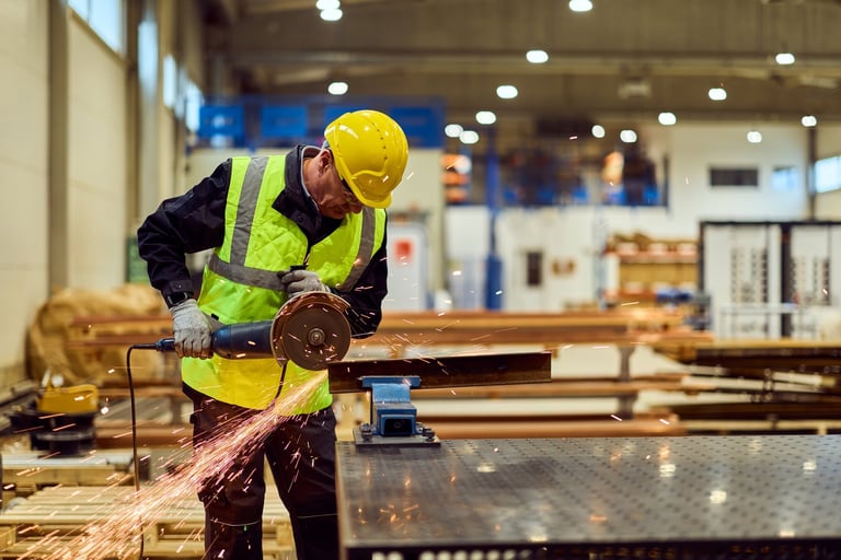 Factory worker using an angle grinder to cut metal amid flying sparks in a busy manufacturing workshop, wearing hard hat, gloves and hi-vis vest — focused on safety, productivity and craft.