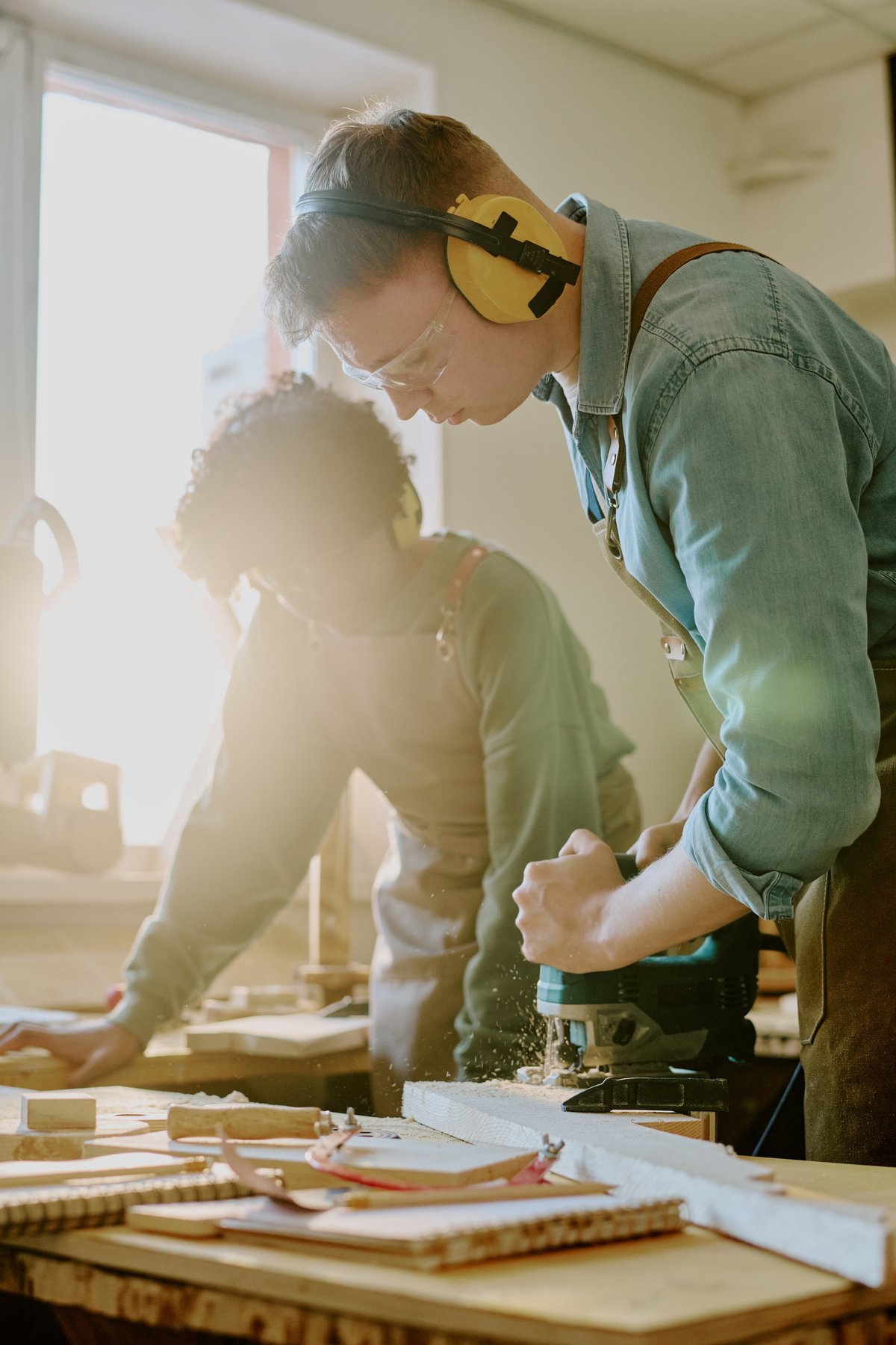 Vertical shot of young adult man operating power router in woodworking class while a woman assists nearby, supporting vocational training and skilled trades education.